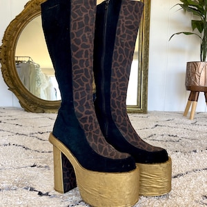 May include: A pair of platform boots with black suede panels and brown animal print accents. The boots have a gold platform sole and heel. The boots are knee-high and have a zipper closure.
