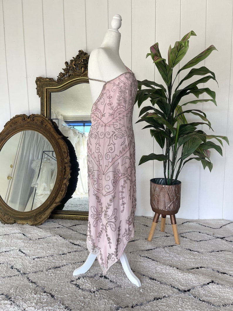 May include: A pink, beaded, sleeveless dress with a plunging neckline and a flowing skirt. The dress is on a white mannequin and has a delicate, intricate design.