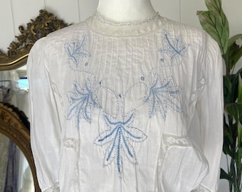 May include: White blouse with blue leaf embroidery. The vintage top features a high neckline, long sleeves, and delicate lace trim. The blouse is displayed on a mannequin, showcasing its intricate details and classic design.