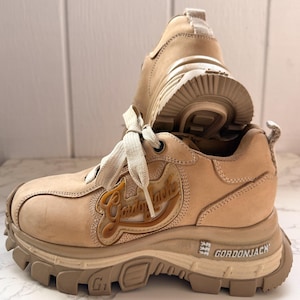 May include: Tan-colored platform sneakers with the brand name "Gordon Jack" in a stylized font. The sneakers have thick, rugged soles and beige laces. The shoes are made of a suede-like material and are designed for casual wear.