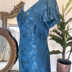 May include: A teal blue maxi dress with a floral pattern and a V-neckline. The dress has a pleated bodice and short, ruffled sleeves.