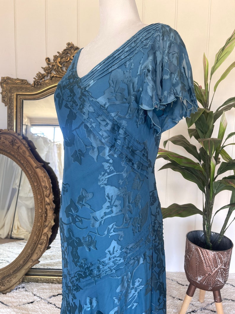 May include: A teal blue maxi dress with a floral pattern and a V-neckline. The dress has a pleated bodice and short, ruffled sleeves.
