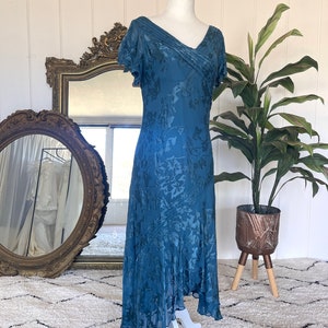 May include: A teal blue silk dress with a floral pattern and a v-neckline. The dress has short sleeves and a flowing skirt.