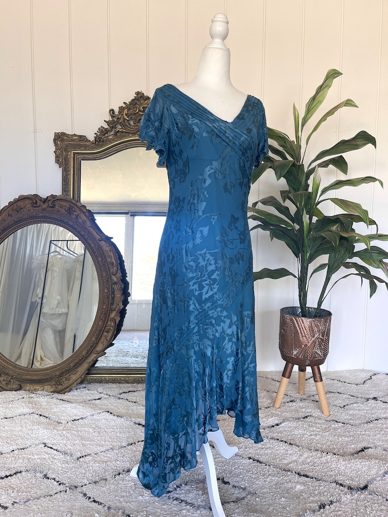May include: A teal blue silk dress with a floral pattern and a v-neckline. The dress has short sleeves and a flowing skirt.