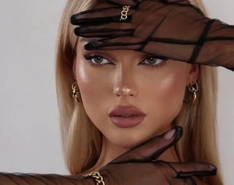 May include: A woman with blonde hair and makeup is wearing black sheer gloves, a gold chain bracelet, and gold hoop earrings. A gold ring is on one finger. The gloves have a grid pattern.