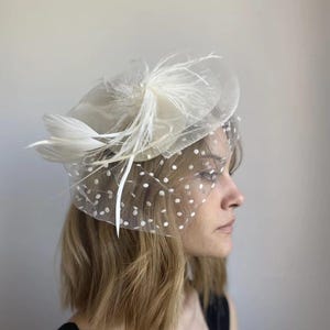 Puede incluir: Un fascinador blanco con velo y detalles de plumas. El tocado presenta una base circular, un velo transparente con lunares blancos y un grupo de plumas blancas. El diseño es elegante y adecuado para ocasiones especiales.