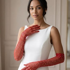 May include: Red fishnet gloves with white rhinestone accents. The gloves are long, extending past the elbows, and are worn with a white dress. The gloves have a delicate, open weave design and are embellished with small, sparkling rhinestones.