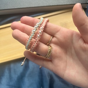 May include: Two macrame bracelets in pastel colours, one light blue and one pink, each adorned with white beads. The bracelets are displayed on a hand, showcasing their delicate design and craftsmanship. The bracelets are a stylish accessory.