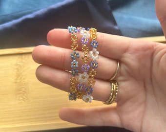 Puede incluir: Tres pulseras de cuentas con un diseño floral. Las pulseras presentan pequeñas cuentas de colores en tonos azules, morados y blancos, ensartadas con metal dorado. Las pulseras se sostienen en una mano, con una bolsa de satén azul en el fondo.