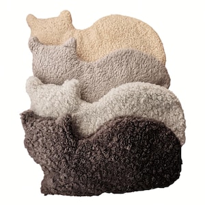 Könnte beinhalten: Ein Stapel von drei Plüschkissen in Katzenform in verschiedenen Brauntönen, Grau und Beige. Die Kissen sind aus einem weichen, flauschigen Material gefertigt und perfekt zum Kuscheln.