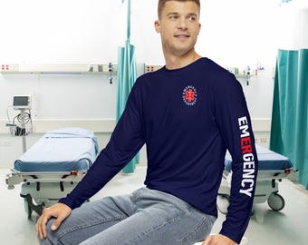 Puede incluir: Camiseta de manga larga azul marino con la palabra "EMERGENCY" en blanco en la manga derecha. La camiseta presenta un emblema de los servicios médicos de emergencia rojo y blanco en el pecho izquierdo. La camiseta se usa con vaqueros azules.