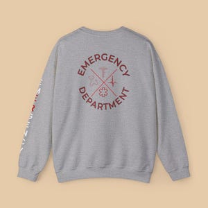 Könnte beinhalten: Ein meliertes graues Sweatshirt mit den roten Worten "Emergency Department", die medizinische Symbole umgeben. Auf dem linken Ärmel steht das Wort "Emergency" vertikal in Weiß. Ein Rundhals-Sweatshirt.