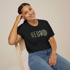 May include: Black t-shirt with the word "NEURO" in a light green font and a brain graphic. The model is wearing blue jeans and sitting on a wooden chair.