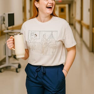 May include: A woman wearing a cream-colored t-shirt with a line drawing of a medical scene, paired with navy blue pants. She holds a cream-colored insulated mug with a handle. The background is a hospital hallway.