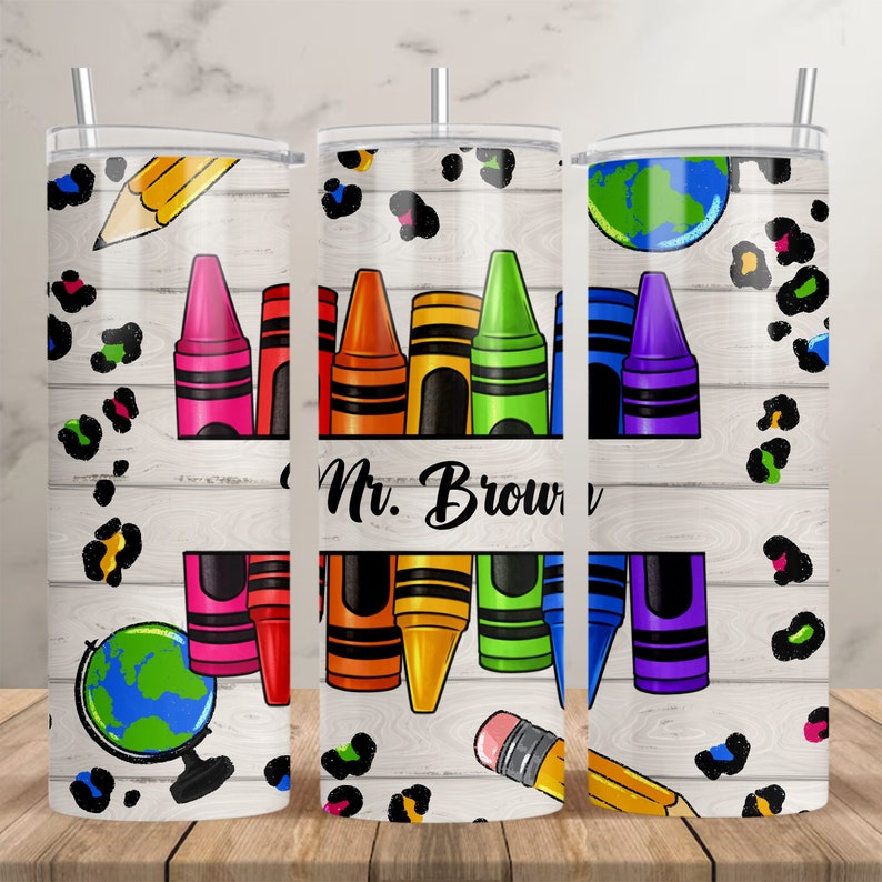 May include: Three stainless steel tumblers with a white woodgrain background. The tumblers feature a design of colorful crayons, a pencil, and a globe. The middle tumbler has the text "Mr. Brown" on it. The tumblers are decorated with a black and white leopard print.