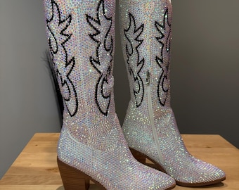 May include: A pair of knee-high western boots completely covered in iridescent, light-reflecting crystals. The boots feature a pointed toe, a stacked wooden heel, and a distinct flame-like pattern outlined in black crystals on the shaft.