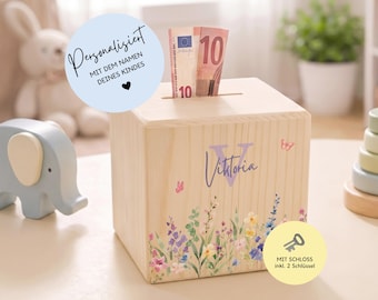 May include: A light-colored wooden money box featuring a slot on top with a ten-euro banknote inserted. The front is decorated with a floral print, a large letter V, and the name Viktoria. It includes a lock and two keys for security.