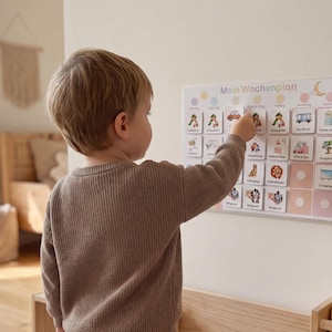 Kleiner Junge steht vor seinem Montessori Wochenplan an der Wand und wählt selbstständig eine Routinekarte für den Tag aus