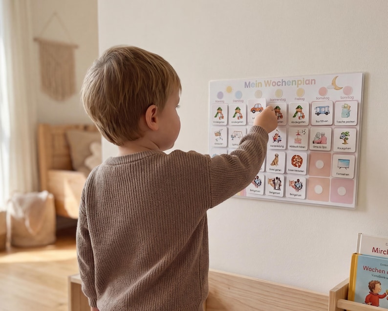 Kleiner Junge steht vor seinem Montessori Wochenplan an der Wand und wählt selbstständig eine Routinekarte für den Tag aus