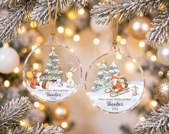 May include: Two clear, round, flat Christmas ornaments with gold hanging strings. Each features a festive illustration of woodland animals, a Christmas tree, and the text "Mein erstes Weihnachten", "Theodor", and "2022" printed on the surface.