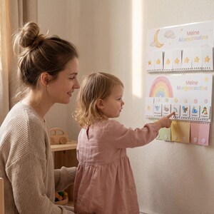Mutter und kleine Tochter im Kinderzimmer vor dem Montessori Routineplaner Regenbogen an der Wand, Kind zeigt auf eine Routinekarte der Morgenroutine