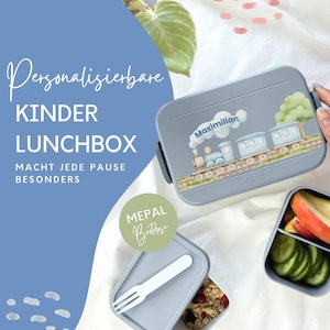 Könnte beinhalten: Eine graue Kinder-Lunchbox mit einem Zugdesign und dem Namen "Maximillian". Die Lunchbox ist geöffnet und zeigt geschnittene Äpfel und Gurken. Ein kleinerer Behälter mit einer Gabel und Müsli ist ebenfalls zu sehen.
