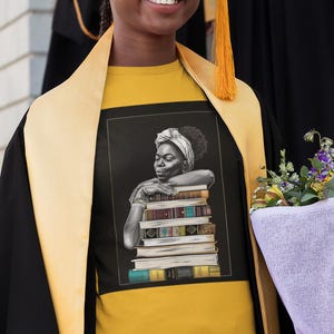 May include: Mustard yellow t-shirt featuring a black and white illustration of a woman leaning on a stack of books. The woman is wearing a headscarf. The person is wearing a graduation gown and holding a bouquet of flowers.