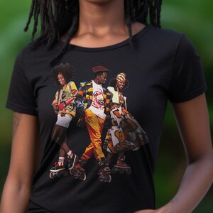 May include: Black t-shirt featuring a colorful graphic of three people roller skating. The graphic includes people wearing vibrant outfits, roller skates, and accessories. The background is a blurred green.