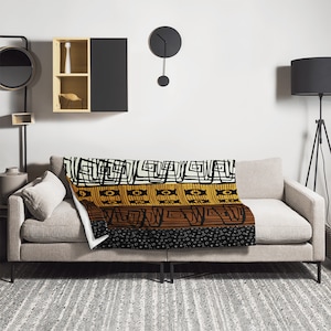 May include: A throw blanket with a geometric pattern in black, white, brown, and yellow. The blanket is folded and placed on a beige sofa.
