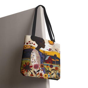 May include: Tote bag featuring a colorful illustration of a woman in profile, set against a backdrop of a field with flowers, a red house, and a yellow sky. The bag has black handles and a cream-colored background.