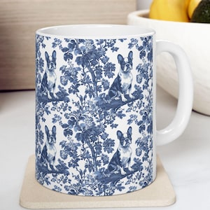 May include: White ceramic mug with a blue and white floral pattern featuring French Bulldogs. The mug has a handle and is sitting on a beige coaster.