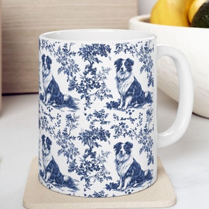 May include: White ceramic mug with a blue and white pattern of border collies and floral designs. The mug has a handle and is sitting on a beige coaster.