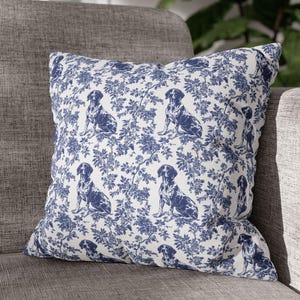 May include: A white cushion with a blue and white pattern of dogs and floral designs. The cushion is sitting on a grey sofa.