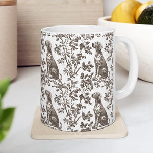 May include: White ceramic mug featuring a repeating pattern of a seated dog and floral elements in brown tones. The mug has a white handle and is placed on a beige coaster. The design gives a vintage, toile-like aesthetic.