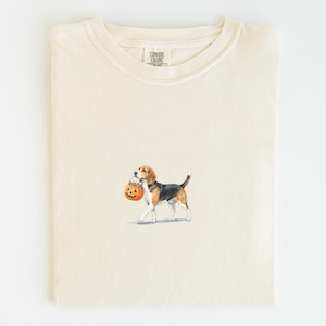Może przedstawiać: Kremowy t-shirt z okrągłym dekoltem, ozdobiony małą grafiką przedstawiającą psa rasy beagle niosącego w pysku wiaderko w kształcie dyni. Wewnętrzna metka na karku zawiera napis Comfort Colors. Koszulka posiada krótkie rękawy i prążkowany kołnierzyk.