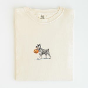 Puede incluir: Una camiseta de cuello redondo color crema con una pequeña ilustración centrada de un perro Schnauzer gris que lleva un pequeño cubo de calabaza naranja en la boca. La camiseta tiene una etiqueta de la marca Comfort Colors en el cuello.