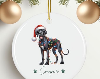 May include: A white ceramic Christmas ornament featuring a black Great Dane dog wearing a Santa hat and Christmas lights. The dog's name, "Cooper", is written in cursive with paw prints on either side. The year "2024" is written below the name. The ornament is hanging from a gold string.