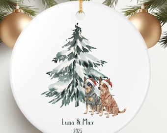 May include: White ceramic Christmas ornament with a watercolor illustration of a snowy evergreen tree and two dogs wearing Santa hats. The ornament has the text "Luna & Max 2025" and a gold hanging cord.