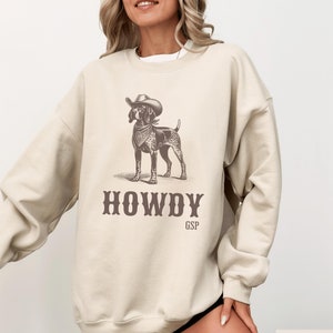 May include: A beige sweatshirt with a graphic print of a dog wearing a cowboy hat and bandana. The text "HOWDY GSP" is printed below the dog.