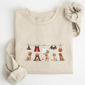 May include: A beige crewneck sweatshirt featuring a graphic of five golden-coloured dogs sitting under a clothesline. The line displays a witch hat, a vampire cape, a ghost sheet, bat wings, and a jack-o'-lantern bucket, with small orange leaves.