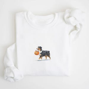 May include: A white crewneck sweatshirt featuring a small, centered graphic of a black, white, and tan Australian Shepherd dog walking while carrying an orange jack-o'-lantern pumpkin bucket filled with colorful candy in its mouth.