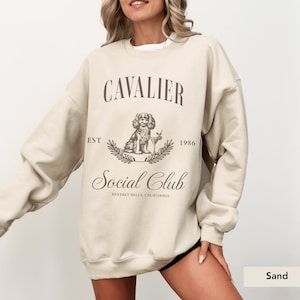 May include: A sand-colored sweatshirt with the text "Cavalier Est 1986 Social Club Beverly Hills, California" and a drawing of a Cavalier King Charles Spaniel holding a martini glass.