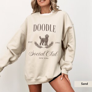 May include: A beige sweatshirt with a doodle dog graphic and the text "Doodle Social Club Est 1986 New York" printed on the front.