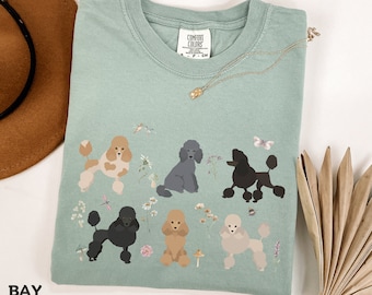 May include: A light green t-shirt with a graphic of six poodle dogs in different colors and poses. The dogs are surrounded by flowers and butterflies. The t-shirt has a small tag that says "Comfort Colors" on the front.