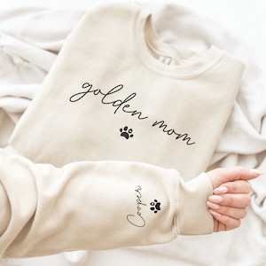 May include: A beige sweatshirt with the text "golden mom" and a paw print on the front. The left sleeve has the text "Cooper" and a paw print.