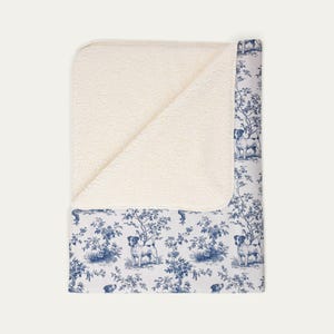 Peut inclure: Un plaid rectangulaire doté d'une face en polaire sherpa blanche et d'une face supérieure à motif toile de Jouy bleu et blanc. L'imprimé présente des illustrations répétitives de chiens debout parmi des arbres et du feuillage sur fond blanc.