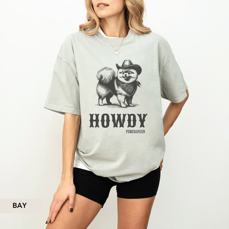 May include: A light green t-shirt featuring a black and white illustration of a dog wearing a cowboy hat and bandana. The text "HOWDY" and "POMERANIAN" is printed below the dog.