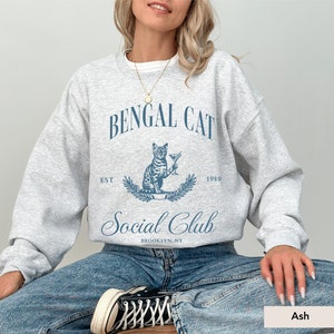 May include: A light grey sweatshirt with a blue graphic that reads "Bengal Cat Social Club" with a cat illustration holding a martini. The sweatshirt also has the text "Est 1989" and "Brooklyn, NY".