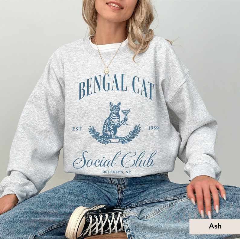 May include: A light grey sweatshirt with a blue graphic that reads "Bengal Cat Social Club" with a cat illustration holding a martini. The sweatshirt also has the text "Est 1989" and "Brooklyn, NY".