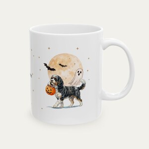 May include: A white ceramic mug featuring a watercolour illustration of a black and white dog carrying a jack-o'-lantern candy bucket. The design includes a large pale moon, a small white ghost, black bats, and tiny stars scattered across the surface.